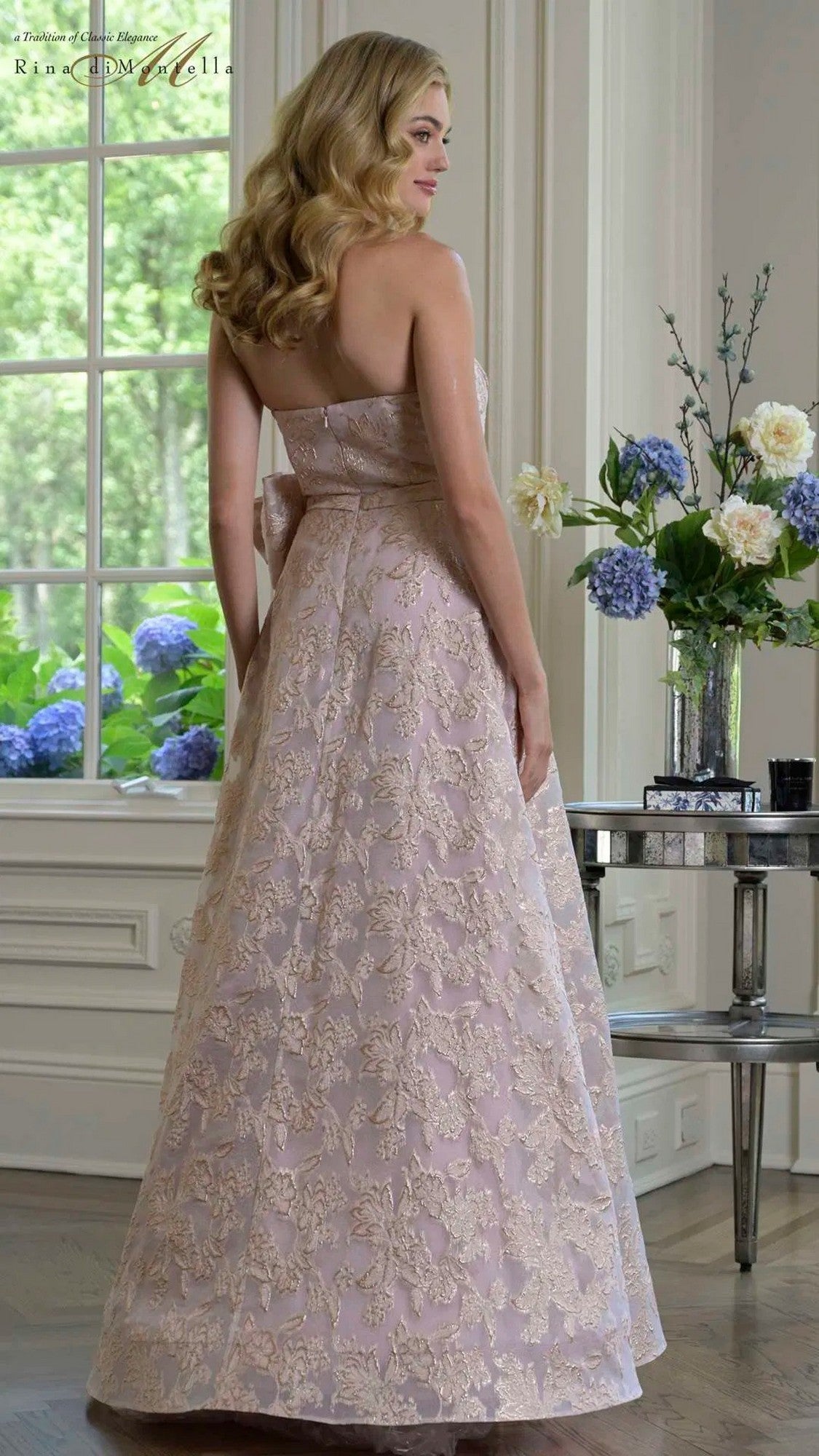 A woman in the Rina Di Montella RD3427 long formal dress, featuring pale pink jacquard fabric and a strapless a-line silhouette, stands in an elegantly decorated room with large windows and floral arrangements.